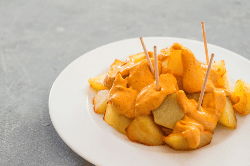 A serving of golden-yellow, roasted or fried potato wedges (Patatas Bravas) is presented on a simple white plate set against a neutral grey background. The potatoes are liberally covered in a thick, creamy, spicy orange-yellow sauce, and several wooden toothpicks are inserted into the pieces, indicating the dish is served as a tapa or appetizer.