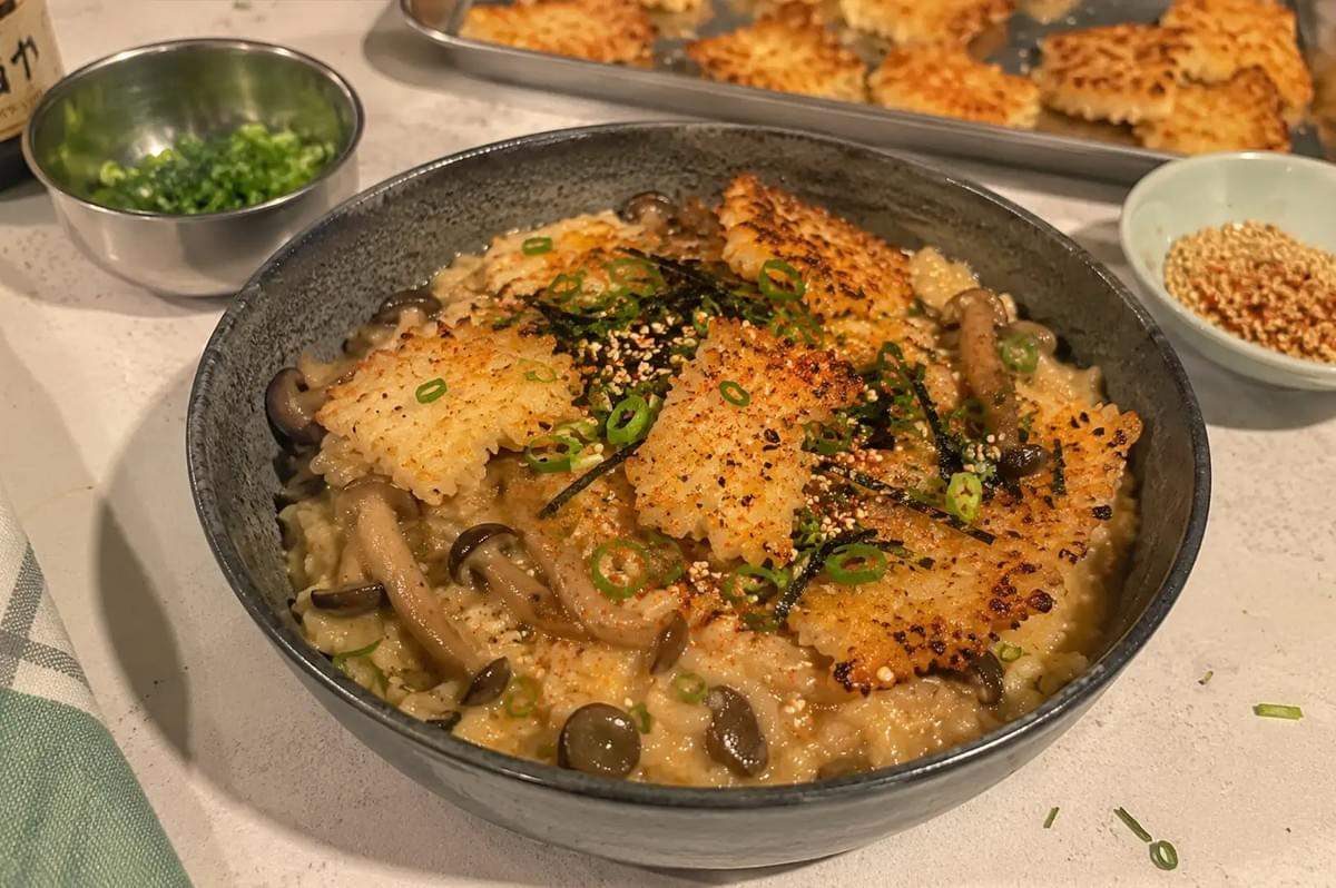 A wide, speckled grey ceramic bowl filled with creamy miso rice and tender shimeji mushrooms, crowned with crisp, golden-brown toasted mochi rice cakes, sliced scallions, shredded nori seaweed, and furikake seasoning, with a baking sheet of crisp rice squares and small prep bowls of green onions and sesame seeds set on a white countertop in the background.