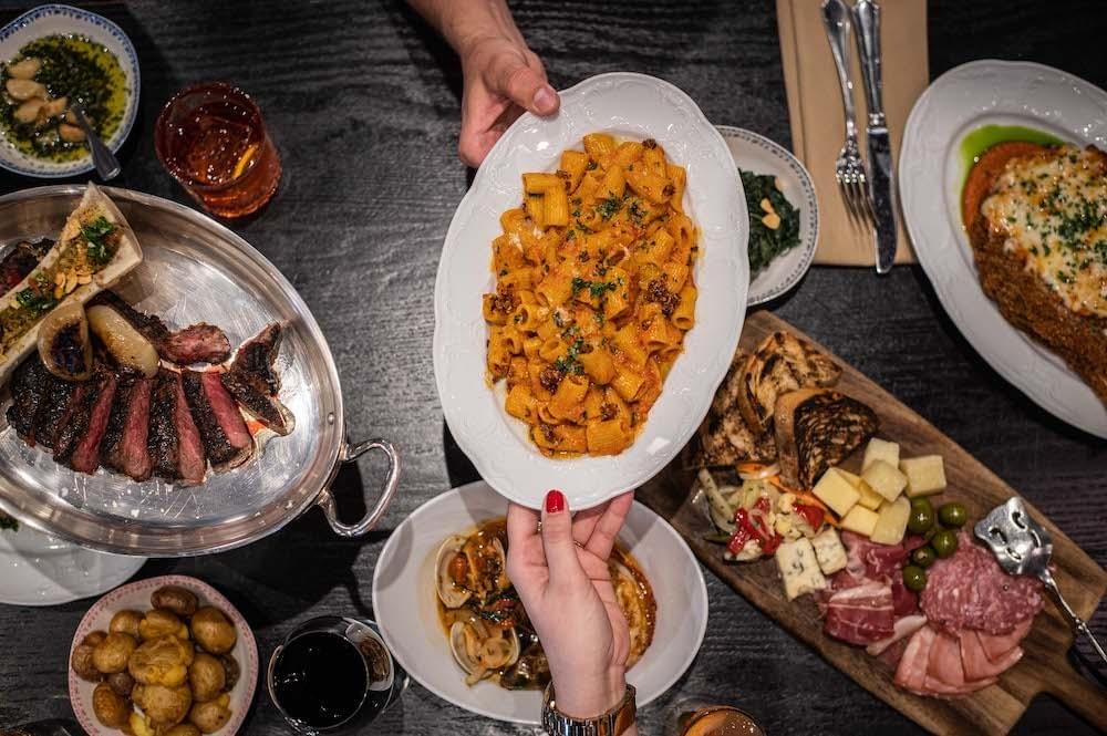 An overhead view captures a dark wooden table lavishly spread with Italian dishes, including sliced steak, a charcuterie board, and roasted potatoes. Two people hold up a central white oval platter filled with rigatoni pasta tossed in a rich orange sauce.