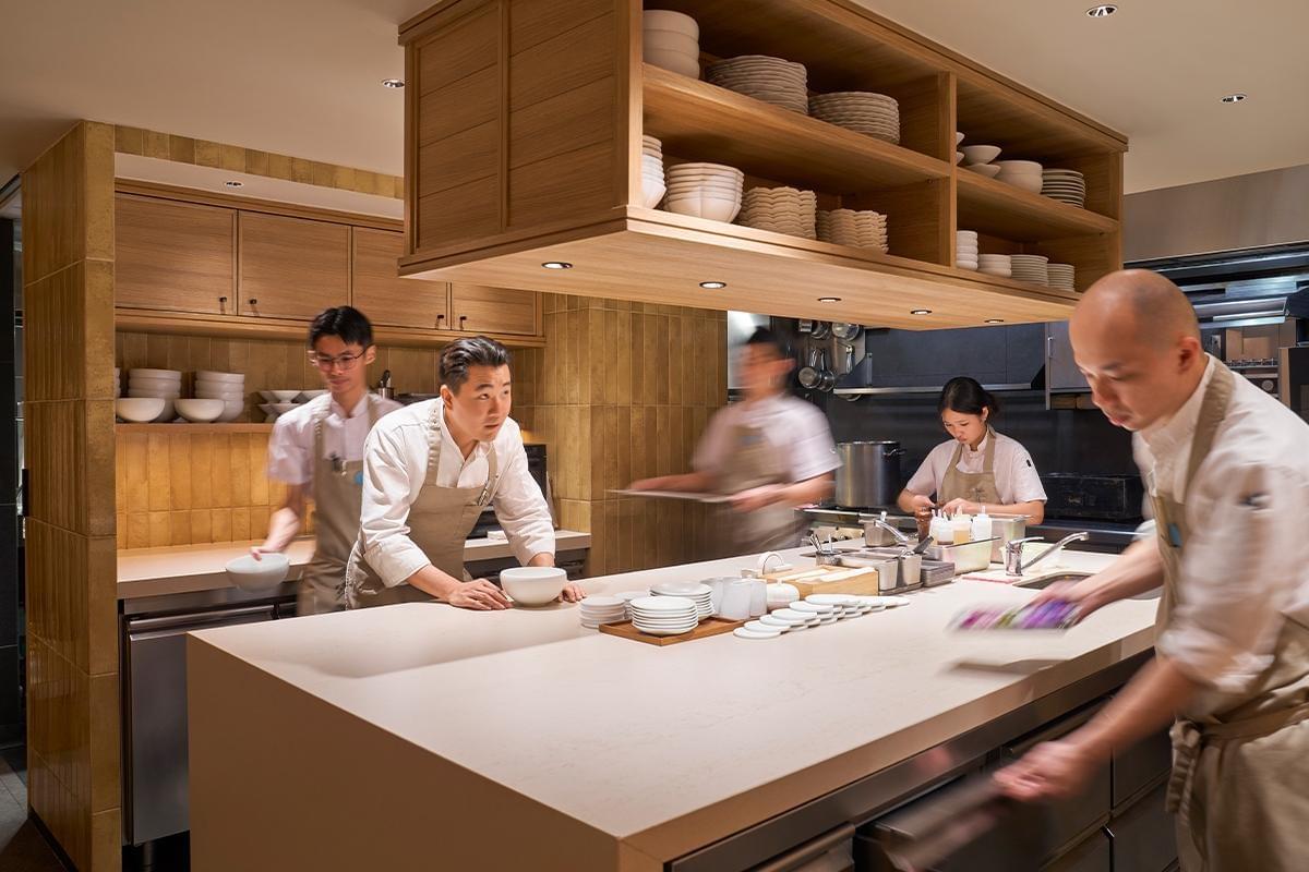 A team of chefs and kitchen staff work busily inside a modern open kitchen with light wooden cabinetry and warm overhead lighting. A main chef leans over the central prep counter alongside several focused assistants moving dynamically through the space.