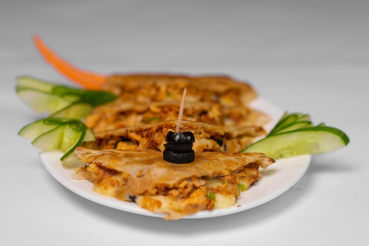 A plate displays several golden-brown quesadilla triangles filled with shredded meat and melted cheese, secured in the center with a toothpick holding black olives. The dish is garnished with fan-sliced green cucumbers and a sliver of carrot on a plain white background.