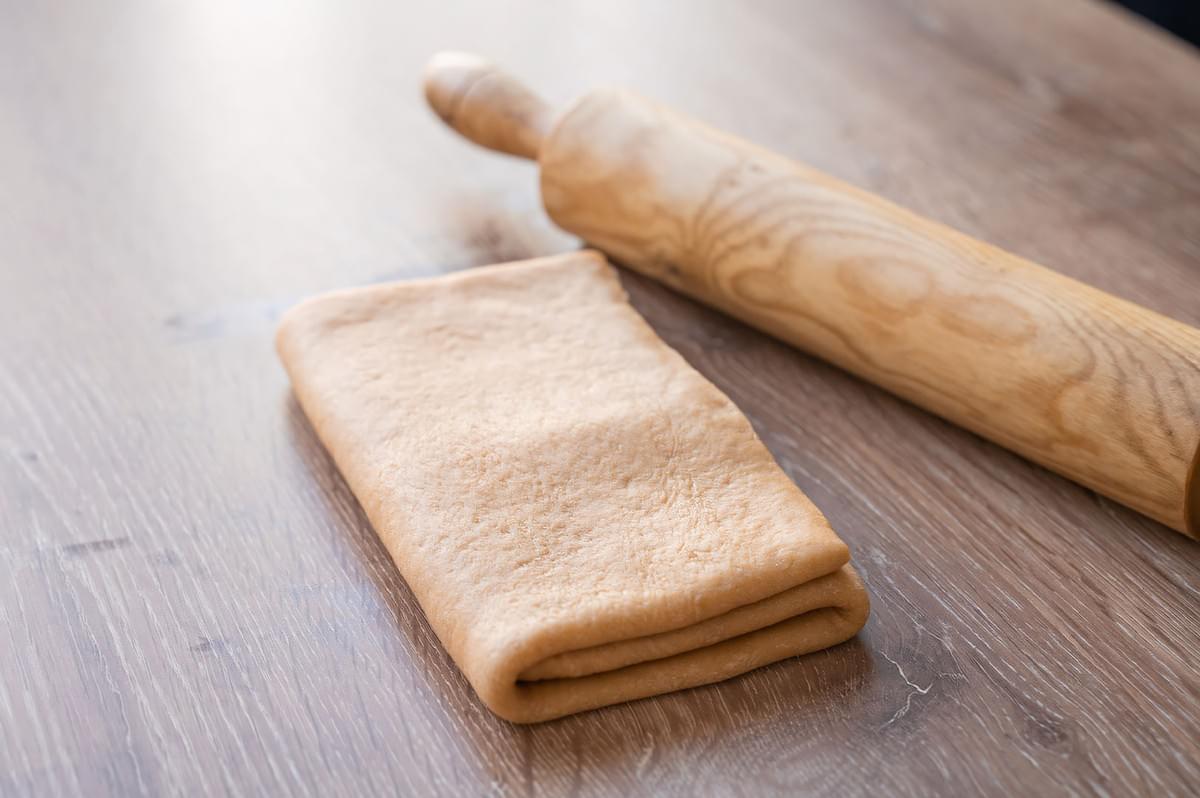 A folded, rectangular block of pastry dough (likely croissant or puff pastry) resting on a wooden countertop next to a wooden rolling pin, showing the critical folding step in the dough lamination process.