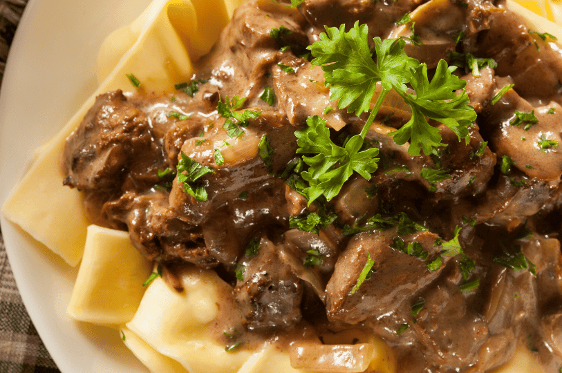 A detailed macro shot highlighting the thick, rich mushroom and beef sauce of a traditional Stroganoff, generously garnished with fresh curly parsley over wide noodles.