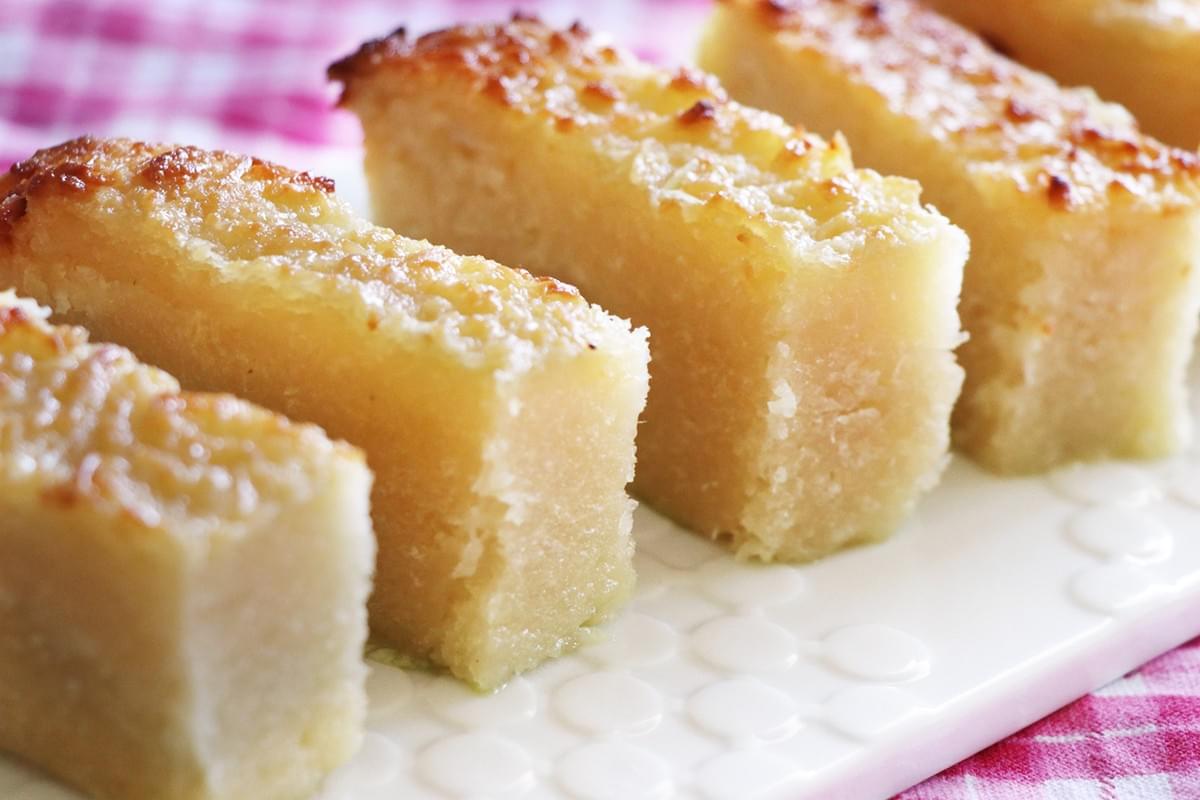 A row of rectangular, golden-brown steamed tapioca cakes sits neatly on a white patterned plate. The treats feature a textured, caramelized top and a dense, translucent center, set against a pink checkered tablecloth.