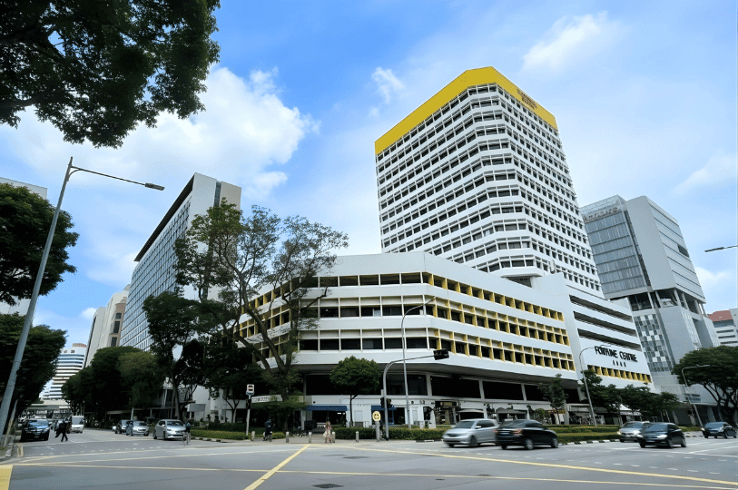 An exterior view of Fortune Centre in Singapore, a multi-story white building with distinct yellow accents under a bright blue sky. The building is a well-known hub for vegetarian and specialty food stalls.
