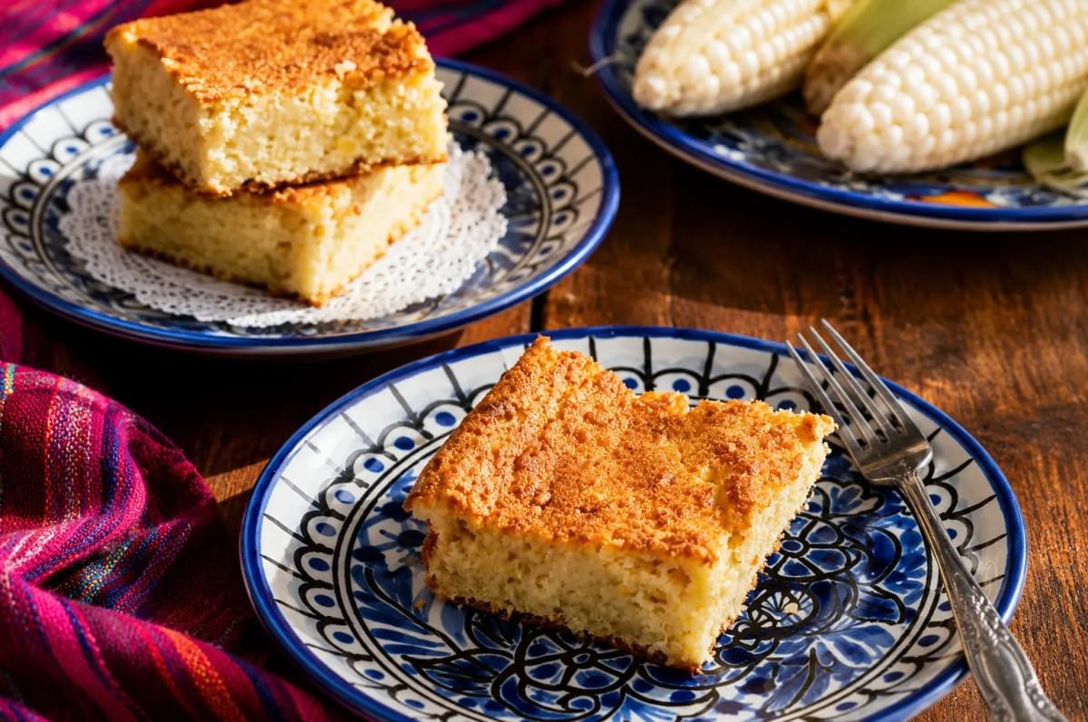 Slice of cornbread on a patterned blue ceramic plate next to fresh ears of corn.