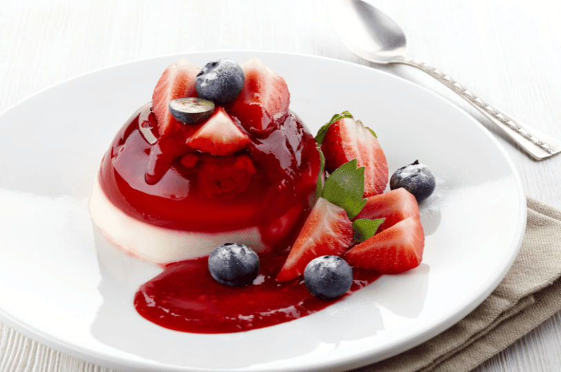 A perfectly set, dome-shaped Panna Cotta covered in a rich berry glaze, surrounded by fresh blueberries, sliced strawberries, and a sprig of mint.