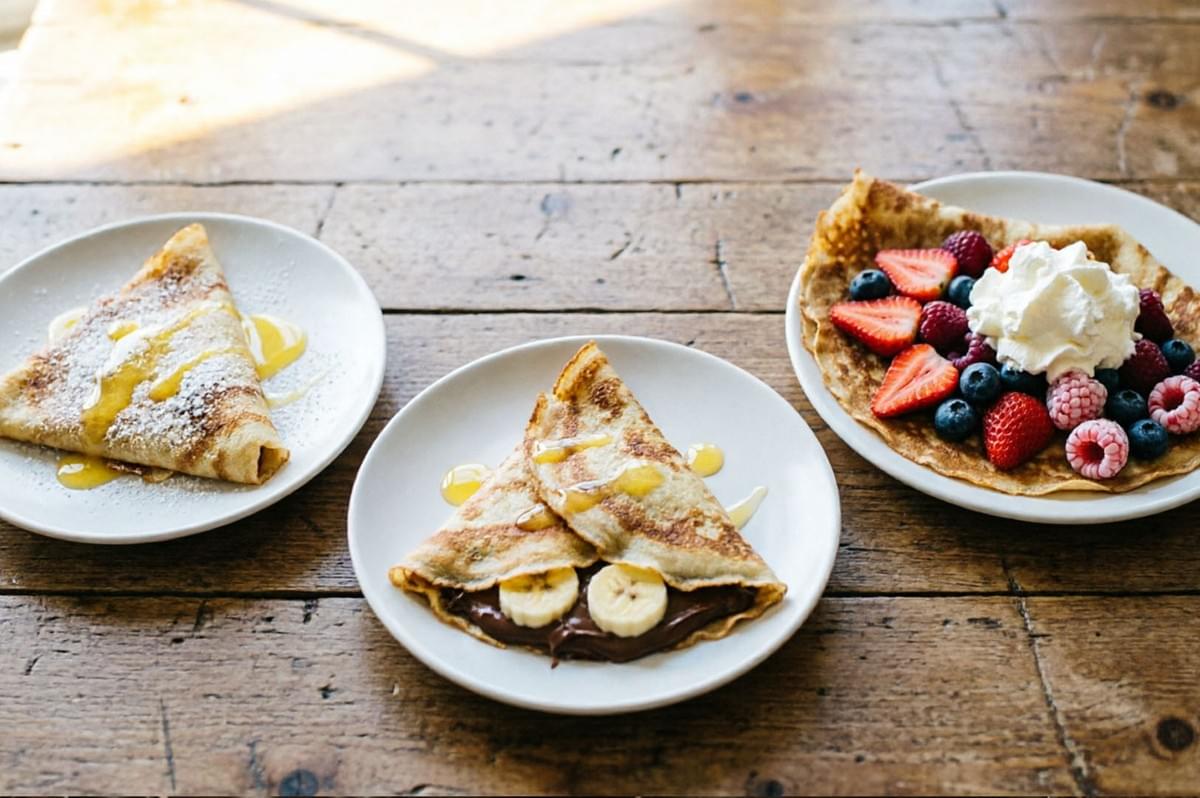 Three sweet crepes on white plates - lemon sugar, Nutella banana, and mixed berries with whipped cream on wooden table