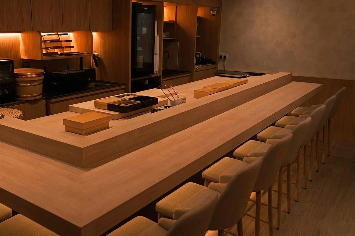 A wide, warm-toned photograph of an empty, upscale sushi counter. The L-shaped bar is made of smooth, light-colored wood, surrounded by minimalist high-back stools with beige upholstery. Behind the counter, the kitchen is organized with wooden cabinetry, recessed amber lighting, and professional tools including a knife rack, a rice barrel (hangiri), and small wooden ingredient boxes (neta-bako).