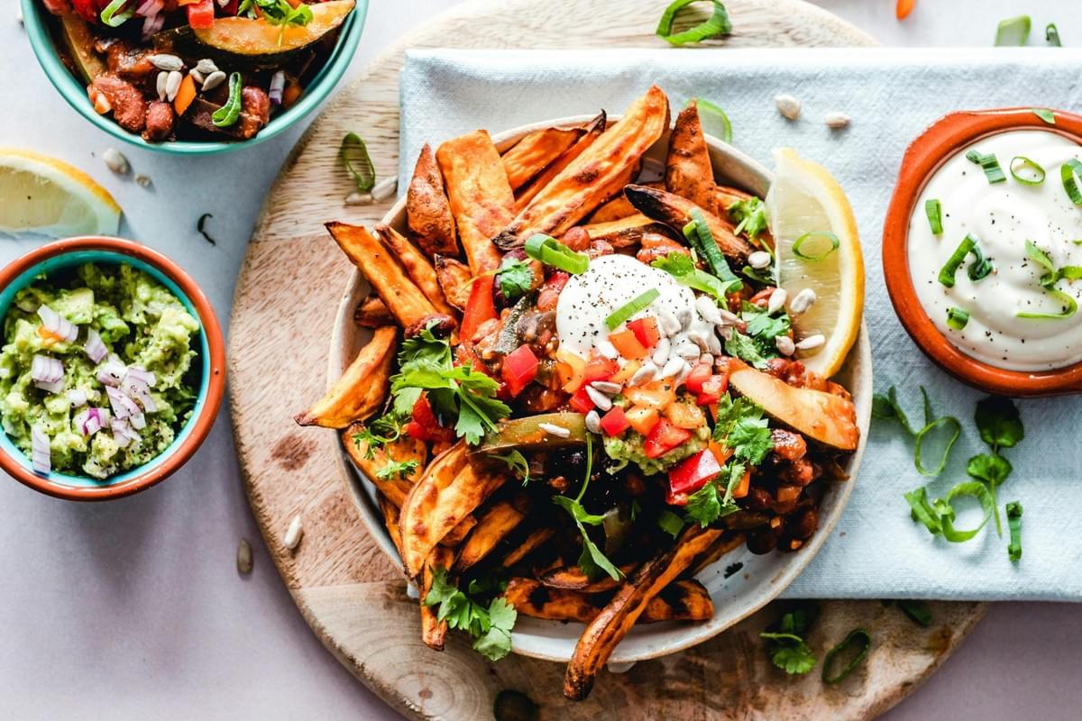 Roasted sweet potato wedges are piled high in a bowl and topped with a savory bean chili, fresh salsa, and a dollop of cream. The vibrant meal is served on a wooden board alongside small dishes of guacamole and a white dipping sauce.