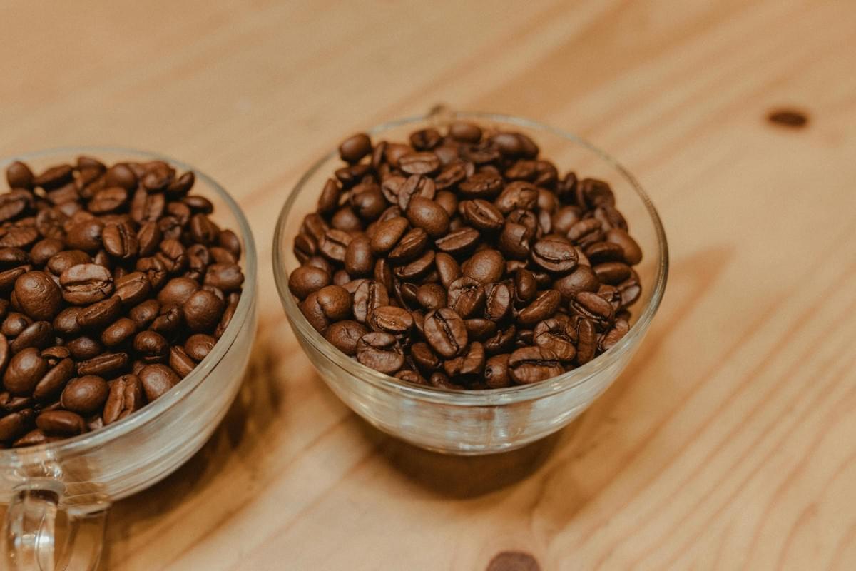 Two clear glass cups filled with roasted coffee beans rest on a smooth, light-colored wooden surface. The warm lighting enhances the rich brown tones of the beans, contrasting them against the natural grain of the table.