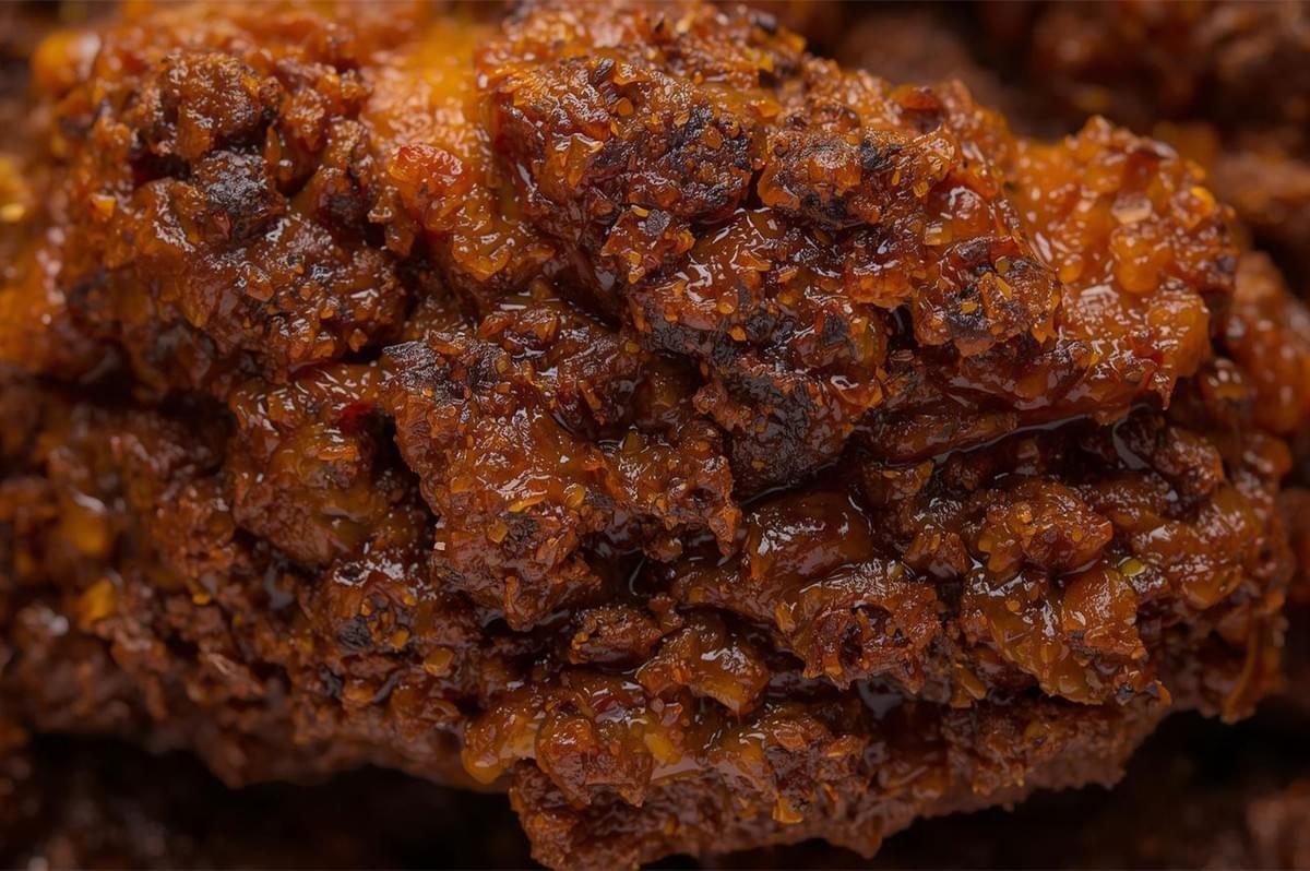 An extreme macro shot of a piece of meat (likely beef) covered in the thick, intensely caramelized, almost dry spice paste of traditional rendang, highlighting its coarse, dark texture.