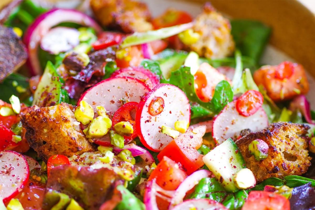 Extreme close-up of a vibrant, colorful salad featuring sliced radishes, chopped tomatoes, red onion rings, small croutons, and chopped pistachios, lightly dusted with spices.