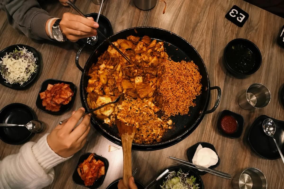 An overhead view of a large circular hot plate filled with dak-galbi, ramen noodles, and fried rice, being shared by people at a wooden restaurant table with various side dishes.