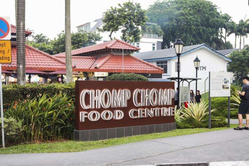 The image features a large brown sign for the Chomp Chomp Food Centre located along a sidewalk in a lush, tropical setting. In the background, there is a traditional building with a red tiled roof and modern white structures nestled among tall palm trees.