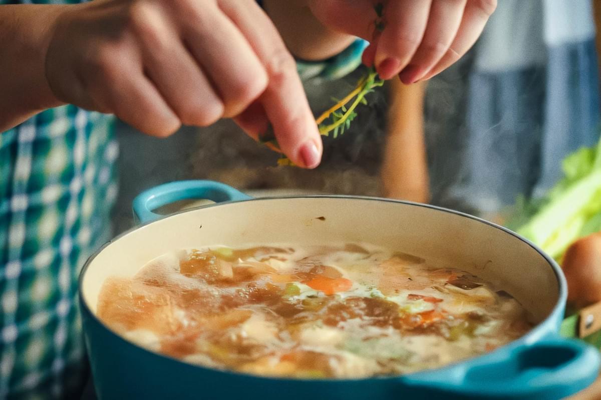 Homemade vegetable soup in blue cast iron pot with carrots, celery, herbs, and meat, person adding fresh thyme sprig.