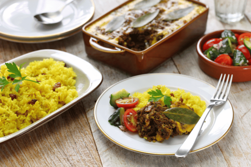 A serving of South African bobotie and yellow rice on a white plate, garnished with a bay leaf and fresh parsley, served alongside a side salad.