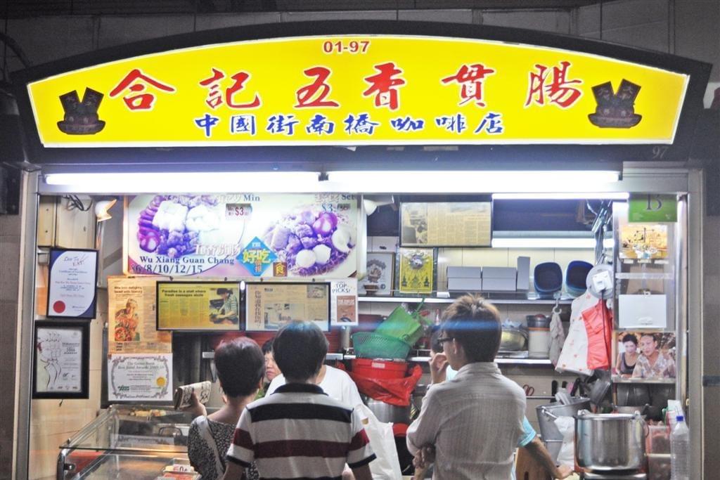 This image shows a bright yellow stall front for Hup Kee Wu Xiang Guan Chang at Maxwell Food Centre, featuring traditional Chinese signage and various food awards. Several customers are standing in front of the counter, waiting for their servings of five-spice meat rolls and other fried delicacies.