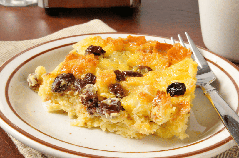 A rustic slice of bread pudding served on a brown-rimmed plate, showing the moist interior and a cluster of dark raisins.
