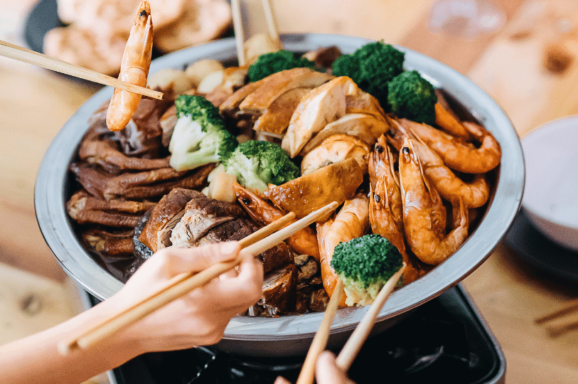A family sharing a communal Pen Cai meal, with multiple pairs of chopsticks picking up prawns, broccoli, and chicken from a large steaming metal basin.