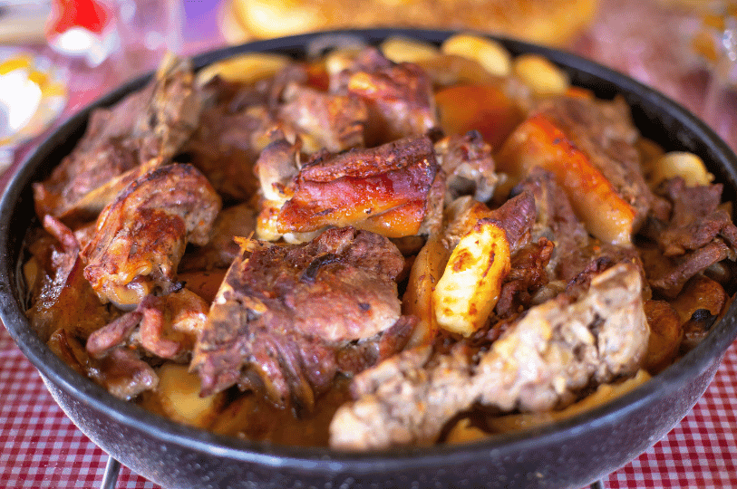 A rustic meat Peka overflowing with large, savory chunks of slow-roasted meat served over sliced potatoes, set on a red and white checkered tablecloth.