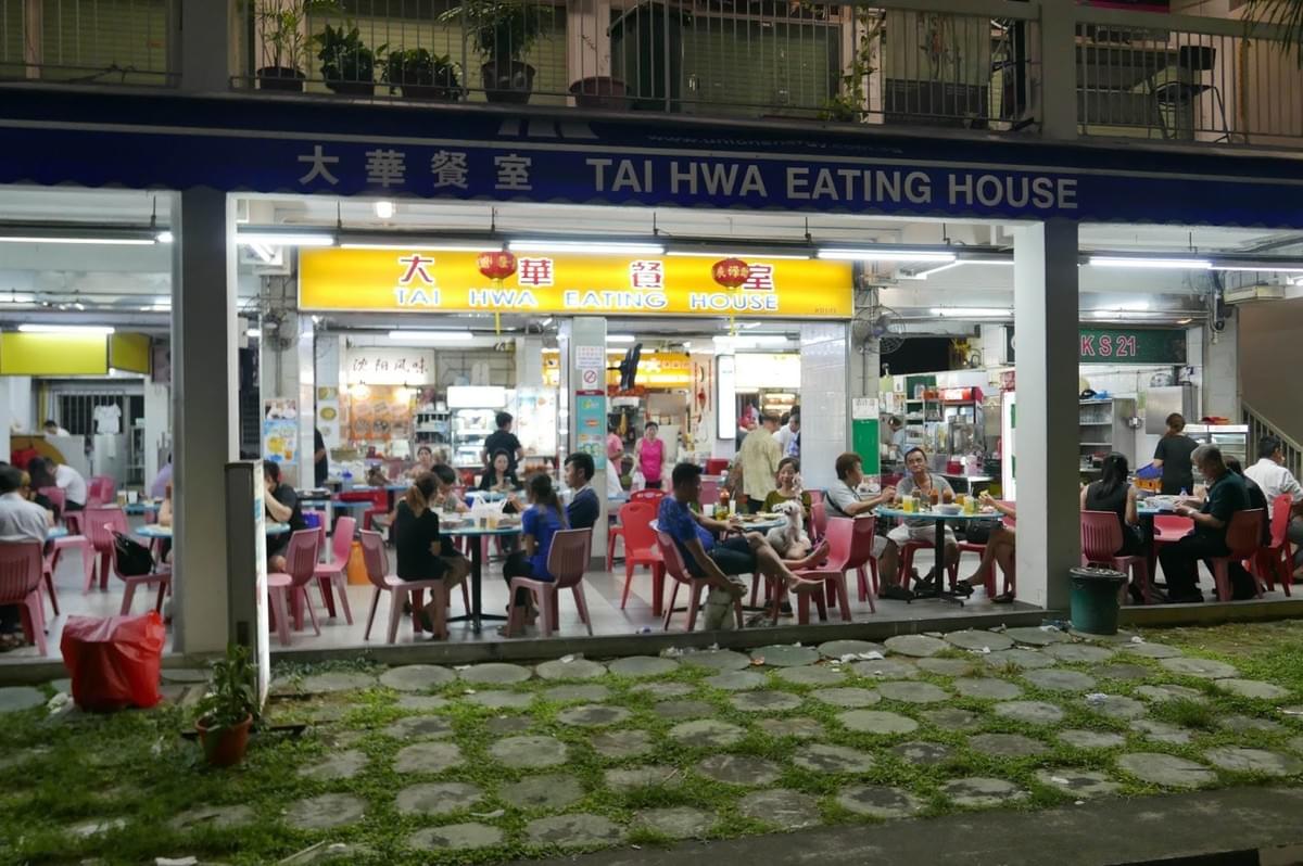 A group of people dining at tables outside Tai Hwa Pork Noodle restaurant, enjoying their meals in a lively atmosphere.