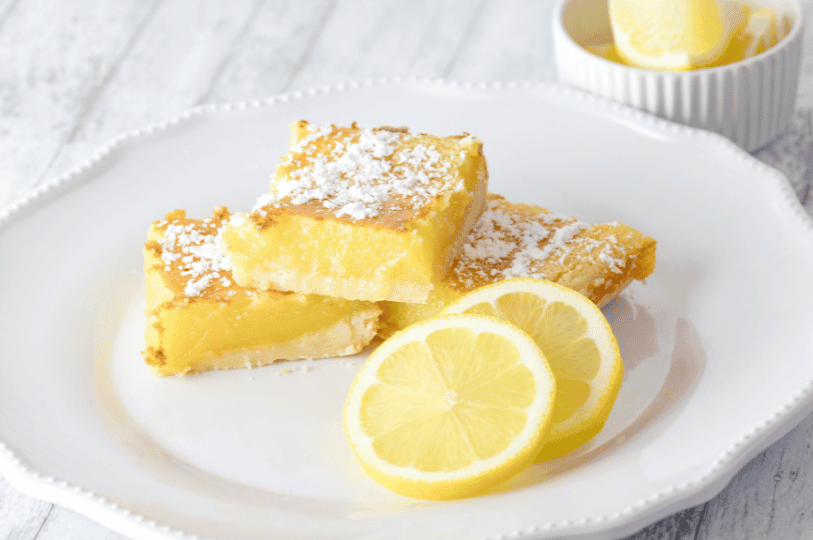 Three lemon bars served elegantly on a white scalloped-edge ceramic plate. The bars are accompanied by two thin lemon slices, with a small bowl of extra lemons in the soft-focus background, creating a bright and airy aesthetic.