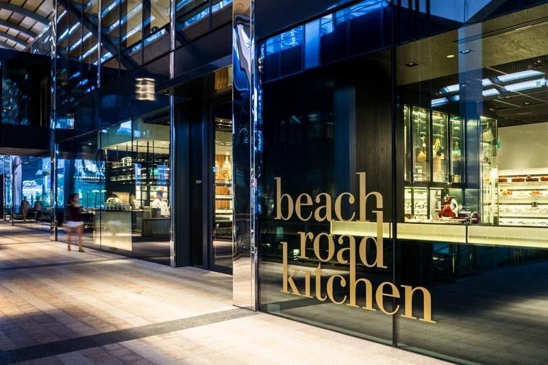 The exterior facade of Beach Road Kitchen features sleek, floor-to-ceiling glass windows displaying the restaurant's name in prominent gold lettering. Warm, modern interior lighting highlights glowing food displays and well-lit kitchen areas inside.