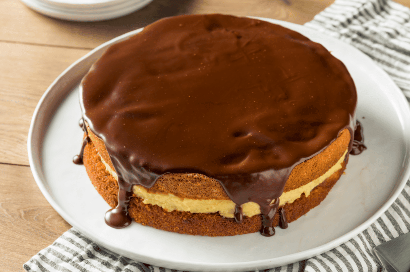 A top-down perspective of a Boston Cream Pie on a white platter, emphasizing the perfectly smooth and reflective surface of the chocolate ganache coating.