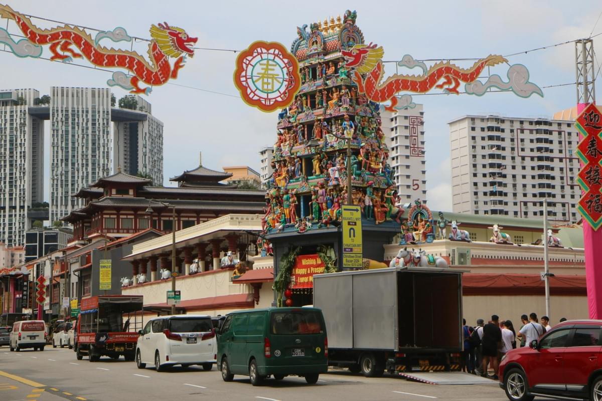 This street scene in Singapore showcases the vibrant Sri Mariamman Temple adorned with colorful sculptures, juxtaposed against a backdrop of modern skyscrapers and traditional Chinese architecture. Festive Lunar New Year decorations, including a large dragon banner, hang over a busy road filled with cars and a delivery truck.