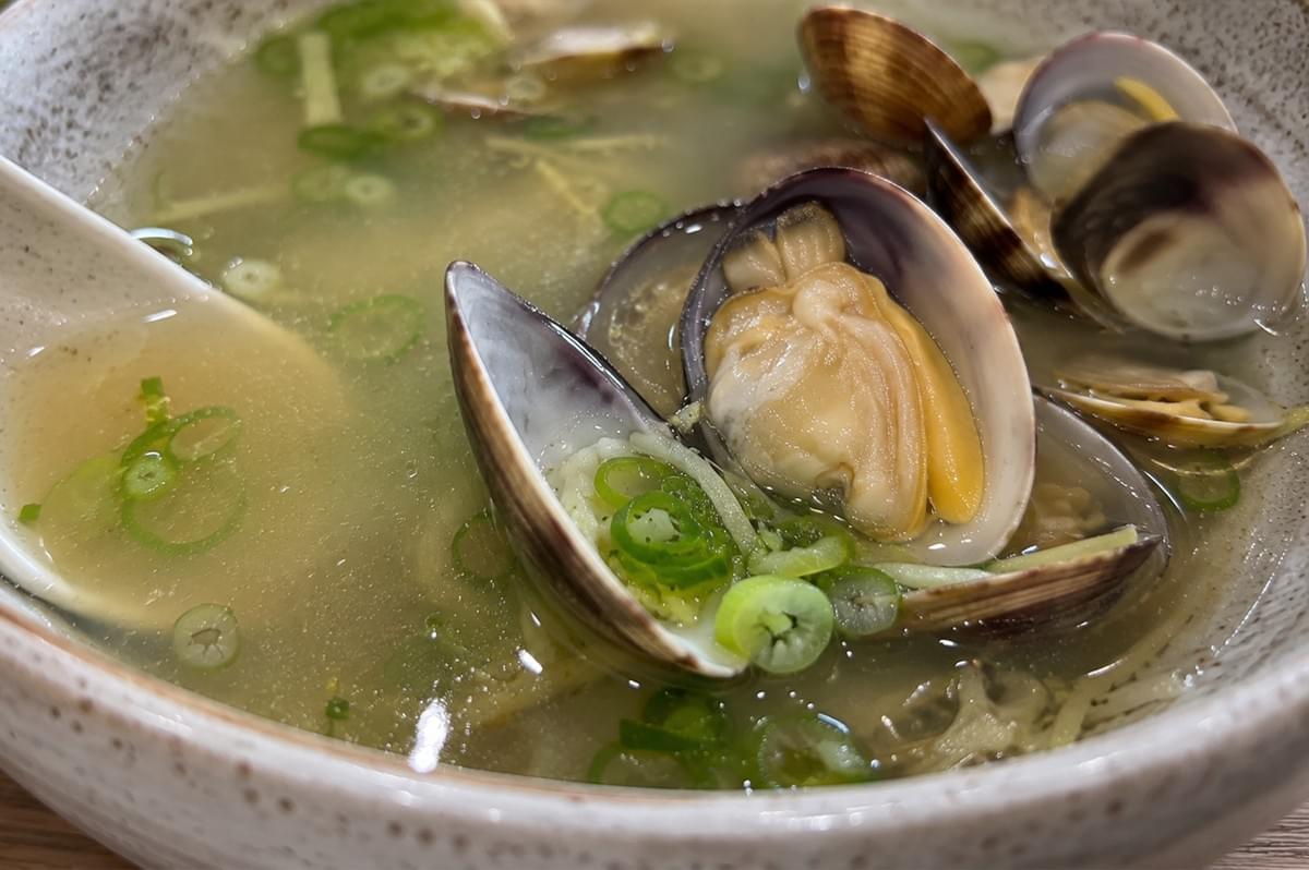 A macro, extreme close-up focuses on a single opened clam sitting in a shallow ceramic bowl of clear broth. The clam meat is plump and yellowish-white, nestled perfectly inside its purplish-brown shell alongside several rings of freshly sliced green scallion. A white ceramic soup spoon is partially visible on the left side dipping into the broth, while other opened clams and fine strands of ginger are softly blurred in the background.