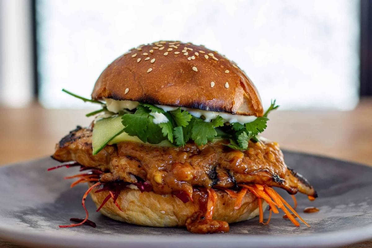 This image features a gourmet burger served on a grey plate, topped with a toasted sesame seed bun and fresh cilantro. The filling includes a grilled protein glazed in a thick sauce, layered with thinly sliced cucumbers and shredded carrots.