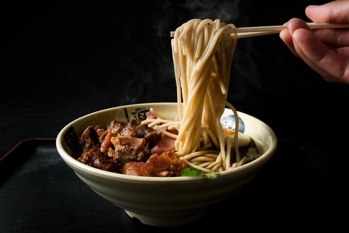 Set against a dramatic dark background, a hand uses chopsticks to lift a large, steaming portion of noodles from a ceramic bowl. The hearty dish features a rich broth topped with generous chunks of braised meat and fresh garnishes.