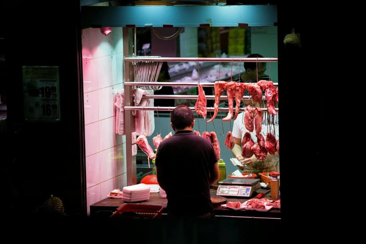 This image captures a butcher stall at a wet market where cuts of fresh meat hang from metal hooks under bright pinkish lighting. A customer stands at the counter while a worker in the background handles transactions near a weighing scale and organized meat trays.