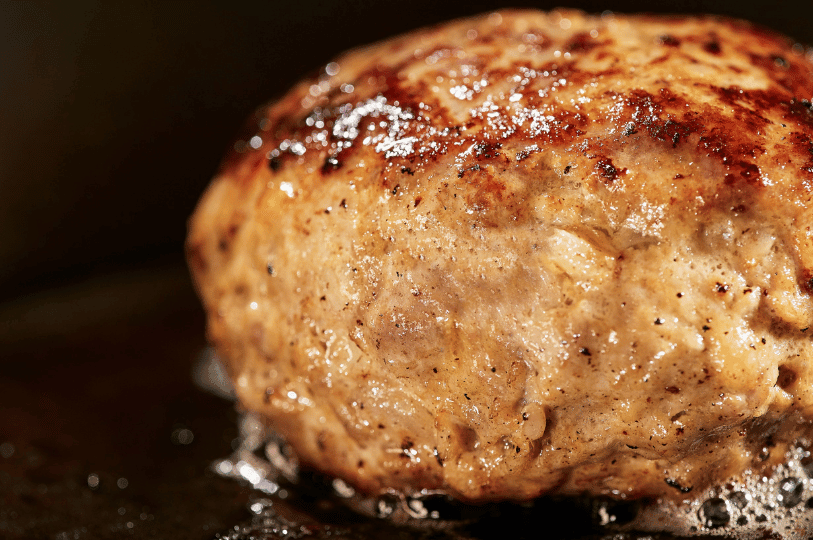 Extreme close-up of a thick burger patty mid-sear, highlighting the textured, caramelized exterior and sizzling rendered fats.