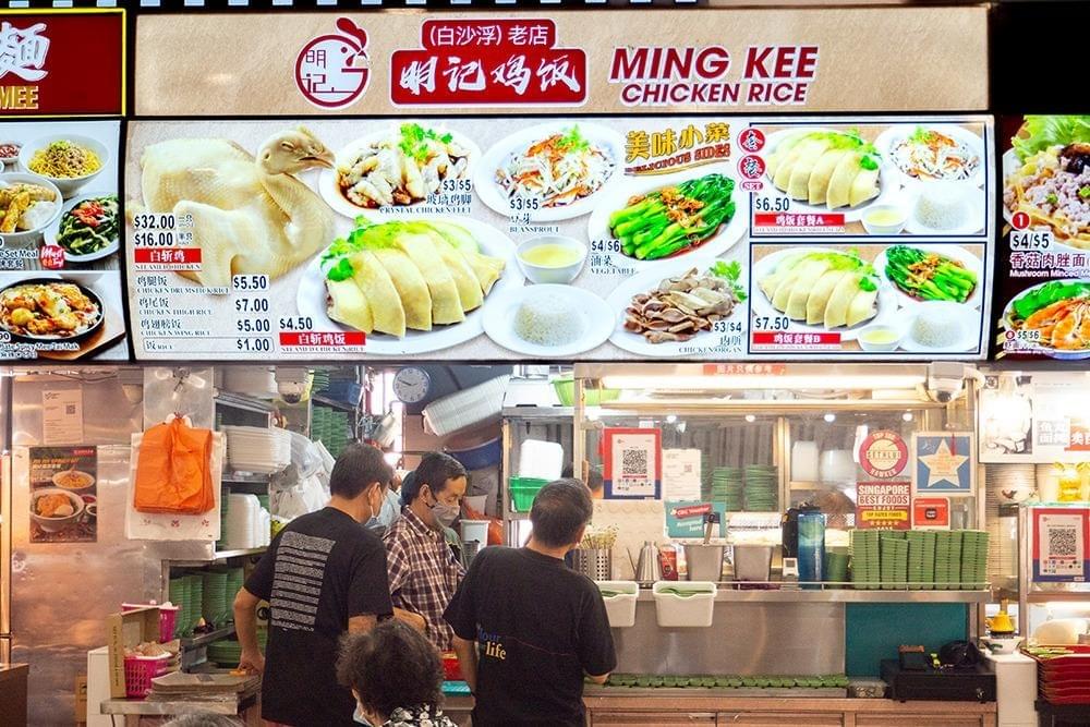  This image shows the Ming Kee Chicken Rice stall, featuring a large, brightly lit menu board that displays various steamed chicken sets, side dishes, and pricing. Below the signage, several staff members in dark t-shirts are busy preparing orders at the stainless steel service counter within a bustling food center environment.