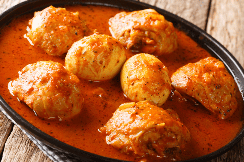 A side-angle close-up of Doro Wat in a black pan, showing tender chicken thighs and hard-boiled eggs simmering in a vibrant orange-red berberé sauce.