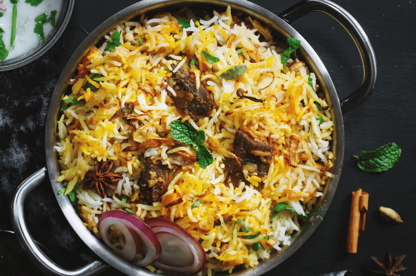 Top-down view of a traditional Afghan Kabuli Pulao in a metal pot, featuring a blend of white and yellow rice, chunks of tender lamb, crispy fried onions, and whole spices like star anise.