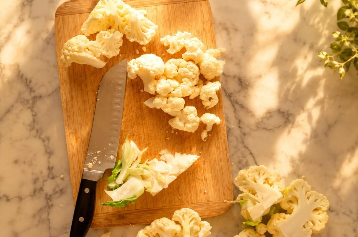 An overhead view of cauliflower florets and a chef's knife on a wooden cutting board, bathed in warm afternoon sunlight.