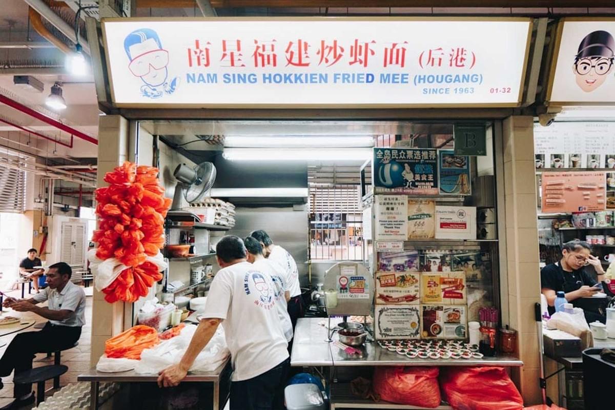 The image depicts the Nam Sing Hokkien Fried Mee hawker stall, which features a prominent white sign with blue and red lettering and a cartoon mascot. Staff members in matching white t-shirts are seen working behind the counter, which is decorated with numerous awards and news clippings.