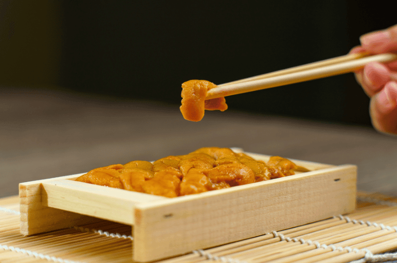 A hand using wooden chopsticks to pick up a piece of fresh sea urchin from a traditional wooden serving box on a bamboo mat.