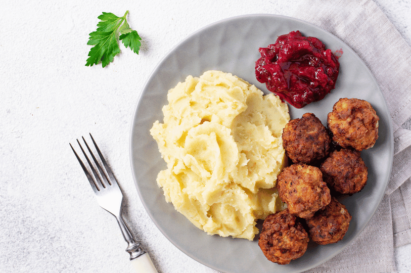 A high-angle, flat-lay shot of a grey plate containing six golden-brown meatballs, a swirl of creamy mashed potatoes, and a portion of textured lingonberry jam, set against a white textured background with a single parsley leaf.