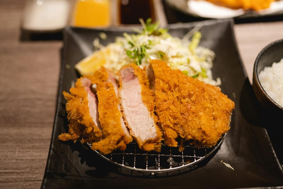 A sliced, golden-brown breaded cutlet rests on a wire rack atop a black plate, showcasing its crispy texture and tender interior. The dish is served with a mound of shredded cabbage salad, a lemon wedge, and a side of white rice to complete the meal.