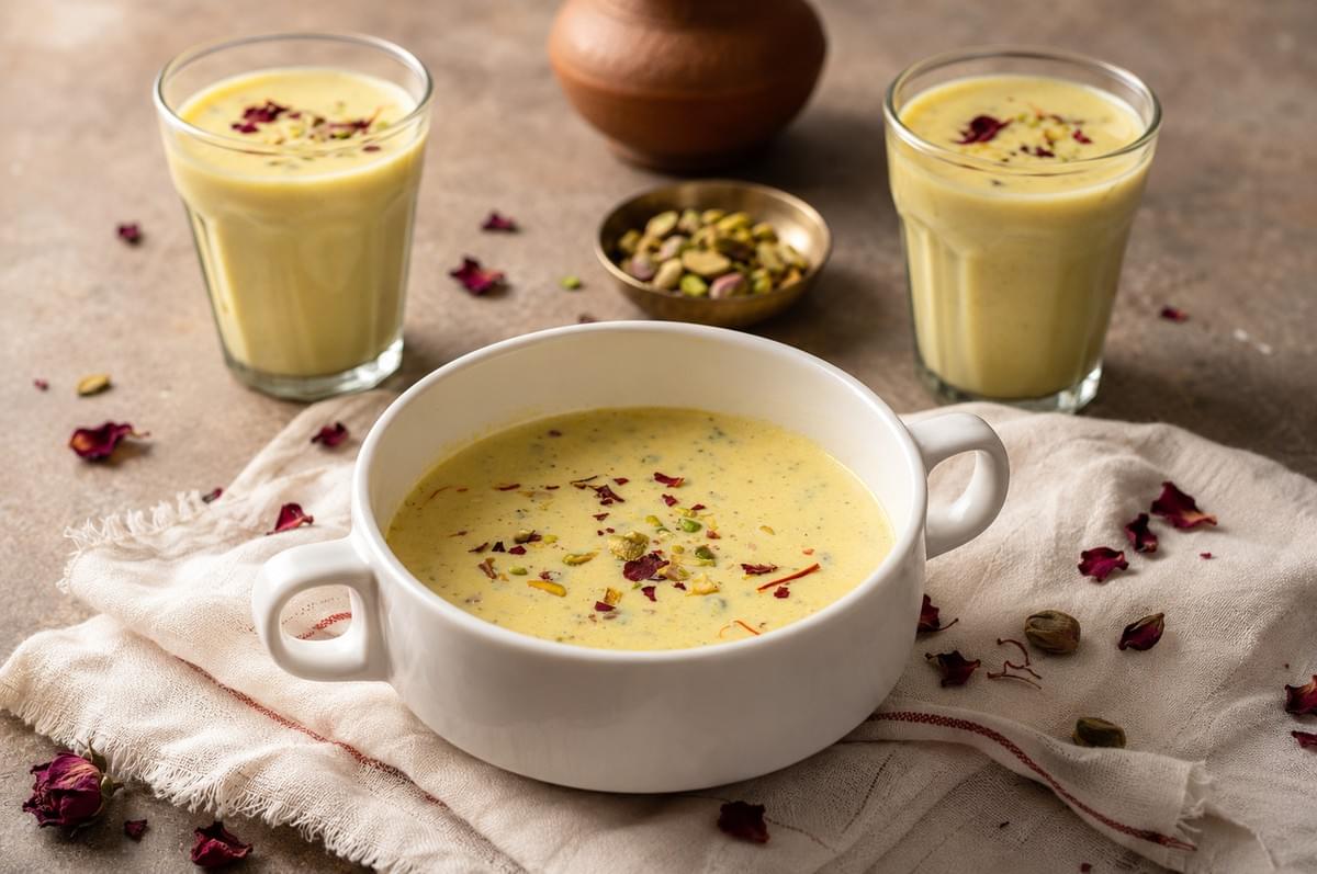 A white bowl and two glasses filled with kheer garnished with pistachios and rose petals.
