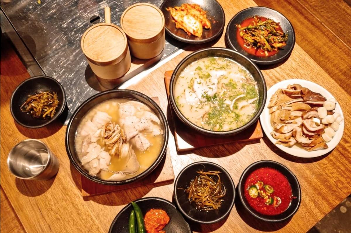 A wooden table with Korean dishes: two steaming soups, various banchan including kimchi, sliced mushrooms, and spicy sauces in small bowls.