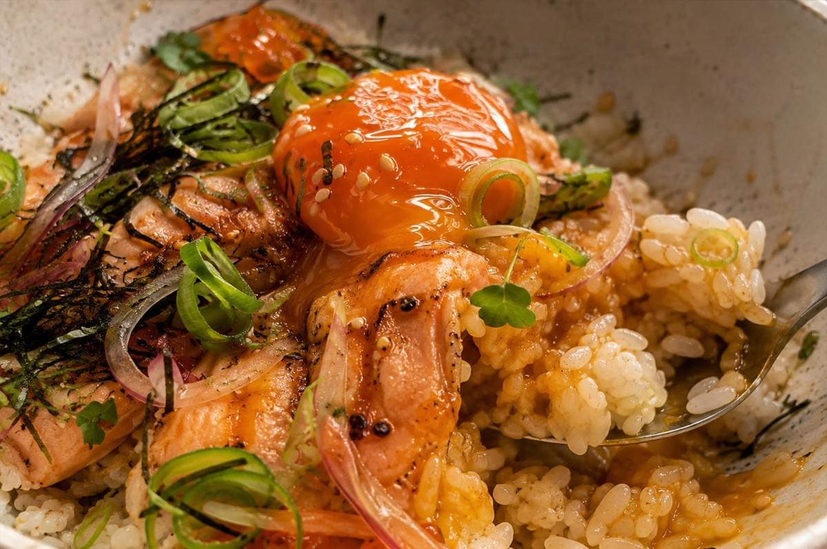 An extreme close-up captures a metal spoon scooping into the chirashi bowl, breaking the glossy soy-cured egg yolk so its rich, golden liquid flows over the seared aburi salmon and warm sushi rice. The detailed shot highlights the torched texture of the salmon, the glistened rice grains coated in yolk sauce, and garnishes of sliced green onions, microgreens, and nori.