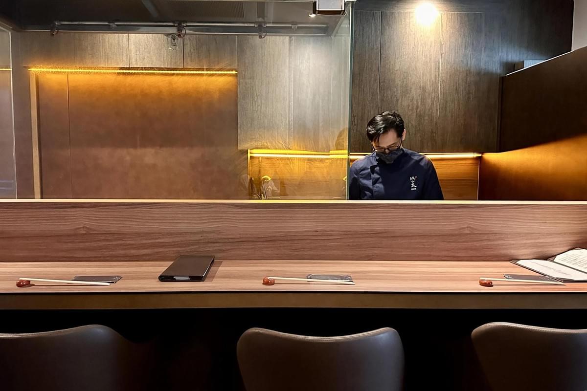 A chef in a dark uniform works behind a sleek wooden counter in a dimly lit, modern restaurant. The foreground features minimalist place settings with chopsticks and leather chairs, all highlighted by warm ambient glow from the back wall.