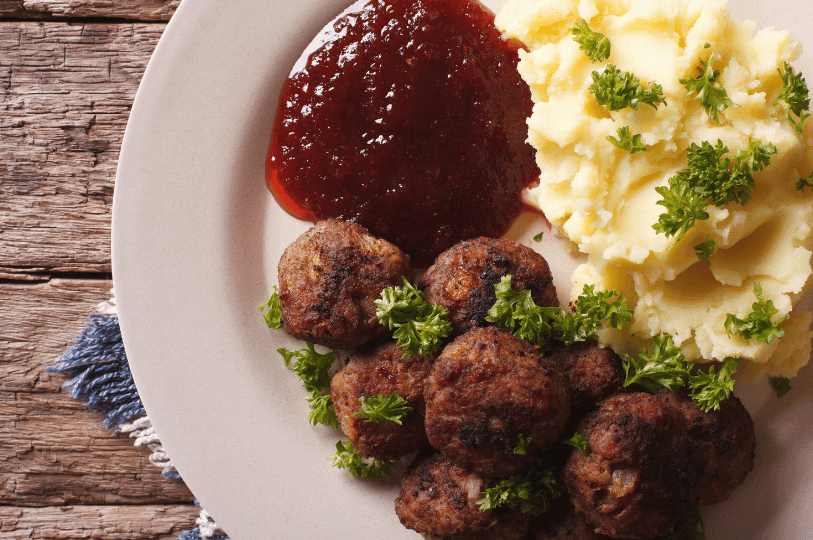 A top-down macro shot of a plate on a rustic wooden table, highlighting the texture of seared meatballs topped with chopped parsley, a smooth pool of lingonberry sauce, and fluffy mashed potatoes.