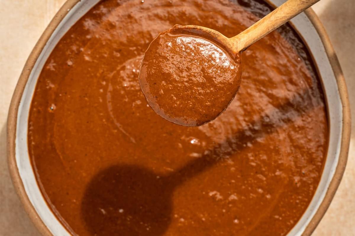 Overhead close-up shot of a wooden spoon lifting a scoop of thick, smooth, reddish-brown mole sauce from a large ceramic bowl, emphasizing the sauce's rich texture.