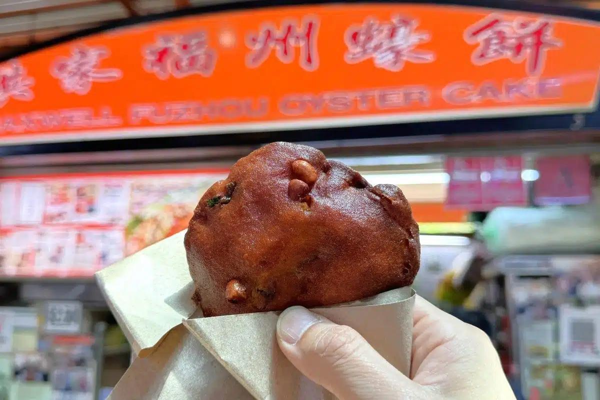 A hand holds a golden-brown, deep-fried Fuzhou oyster cake in a paper wrapper, with several peanuts embedded in its crispy surface. In the background, the bright orange signage of a hawker stall at Maxwell Food Centre is visible.